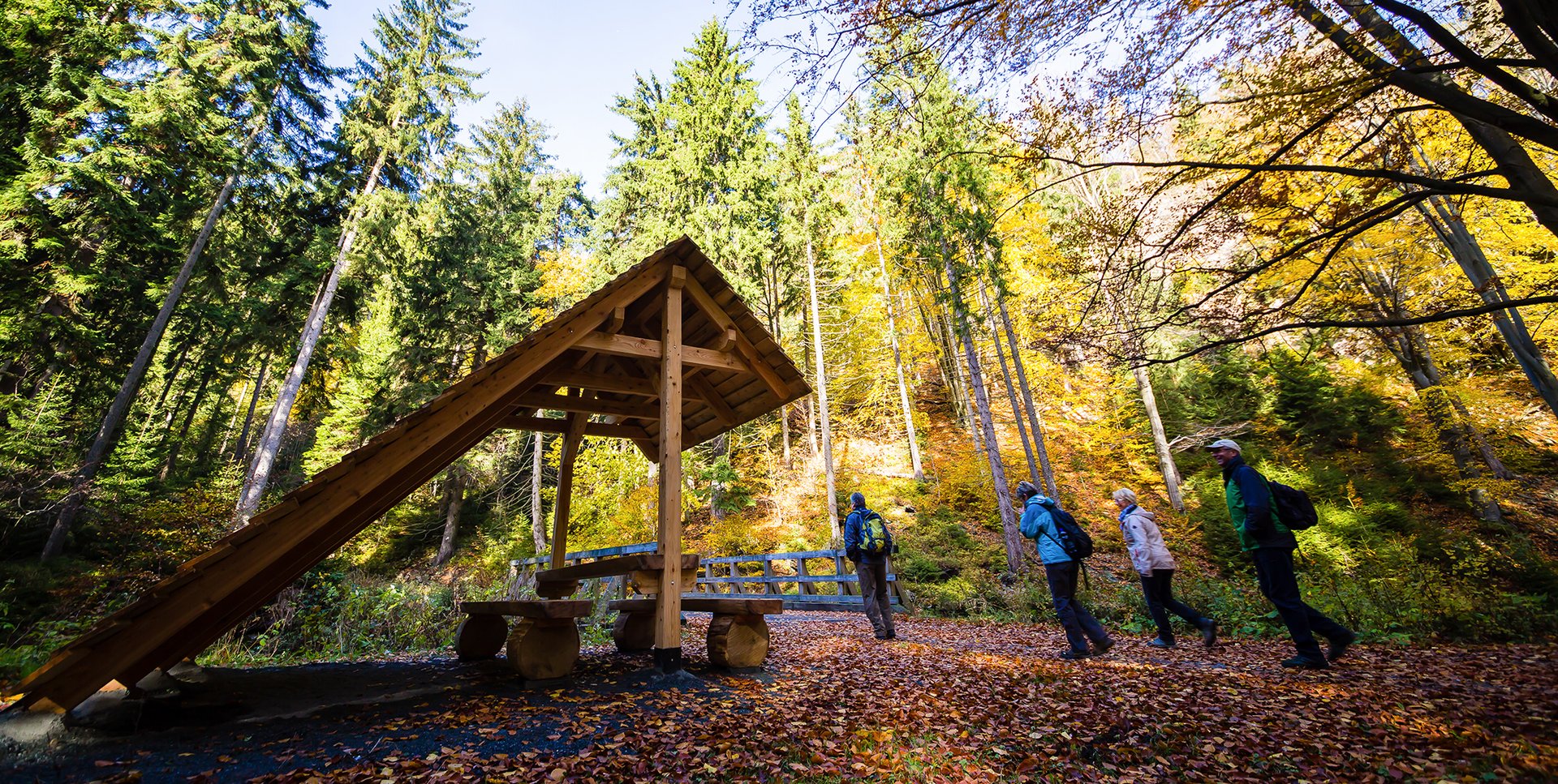 Wanderweg mit Schutzhütte. Links befindet sich eine Schutzhütte und rechts davon befinden sich 4 Menschen. Im Hintergrund befinden sich viele Bäume.