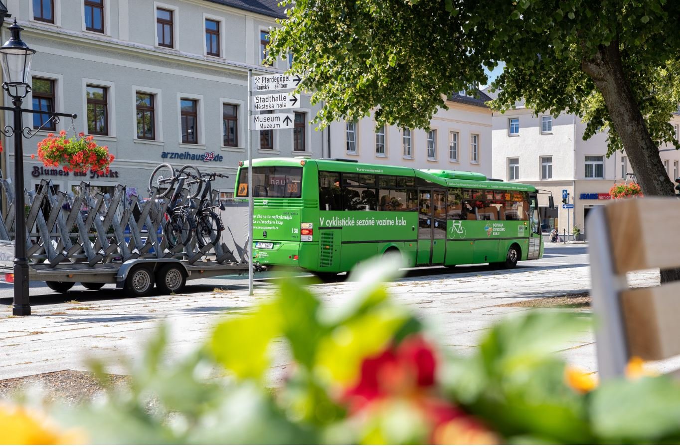 Grenzbus. Im Vordergrund befinden sich Blumen. Im Hintergrund befindet sich ein grüner Bus, auch Grenzbus genannt, mit einem Fahrradanhänger. Der Bus befindet sich an einer Bushaltestelle.