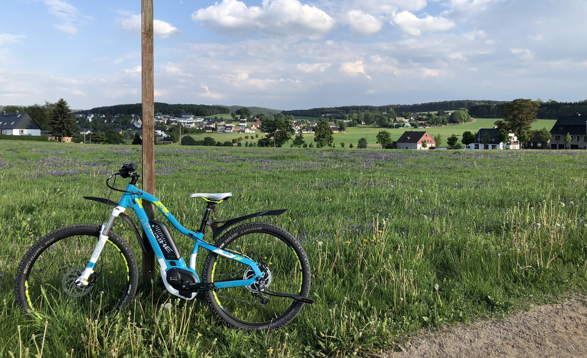 Fahrrad mit Blick auf Gebirge. Im Vordergrund befindet sich ein blau/weißes Fahrrad. Und im Hintergrund befindet sich eine Wiese mit Häusern-